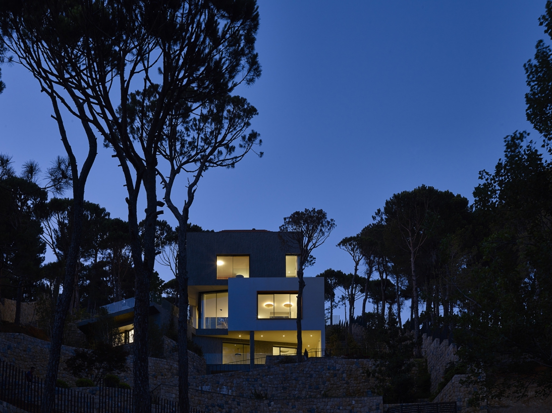 Nocturnal view of the southern elevation of the building, surrounded by the vegetation. The volumes arranged on top of each other, suspended on the landscaped slope. The top volume has a pitched rood and is covered in grey stone, the intermediate volume finished in white paint and is advanced.